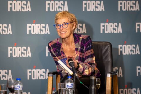 Jane McAlevey sits holding her book and in front of a microphone in front of a background with "Fórsa" written on it.